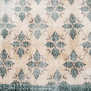 May include: A woven textile rug featuring a repeating pattern of dark teal floral motifs set against a light beige background. The design includes a solid dark teal border along the left and bottom edges, with a scalloped edge detail.