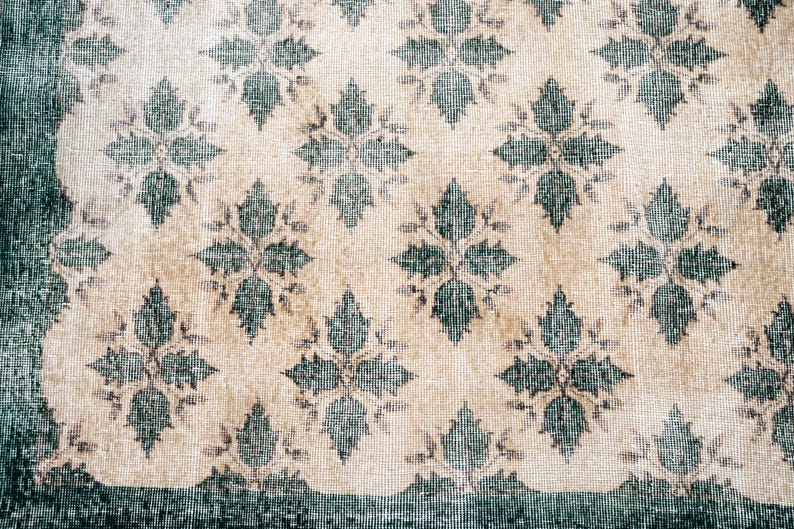 May include: A woven textile rug featuring a repeating pattern of dark teal floral motifs set against a light beige background. The design includes a solid dark teal border along the left and bottom edges, with a scalloped edge detail.
