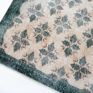 May include: A rectangular rug featuring a beige center field decorated with a repeating dark teal floral pattern. The rug has a wide, solid dark teal border along the edges and a short, light-colored fringe along the visible corner edge.