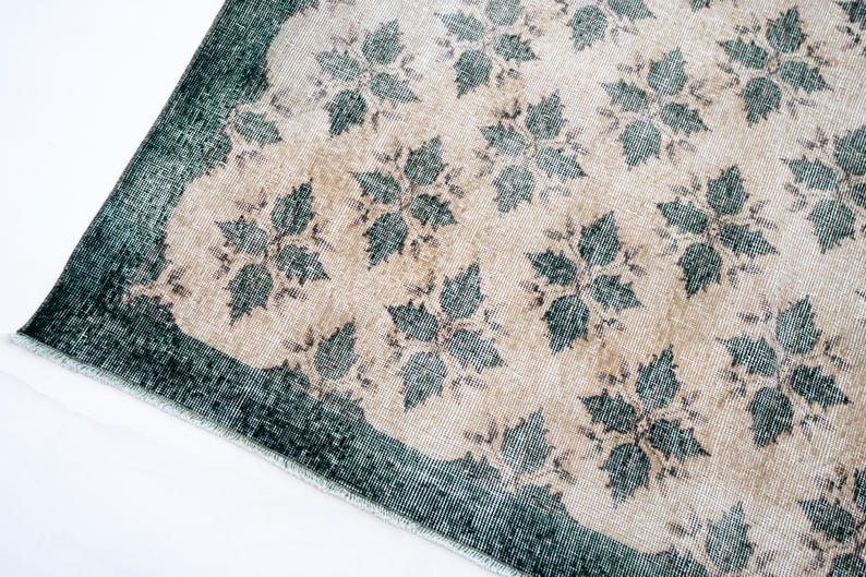 May include: A rectangular rug featuring a beige center field decorated with a repeating dark teal floral pattern. The rug has a wide, solid dark teal border along the edges and a short, light-colored fringe along the visible corner edge.
