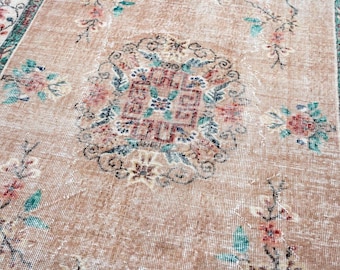 May include: A rectangular rug featuring a tan base color with a central medallion containing a red geometric pattern surrounded by floral motifs in red, green, and white. The border displays a repeating pattern in green, red, and white.