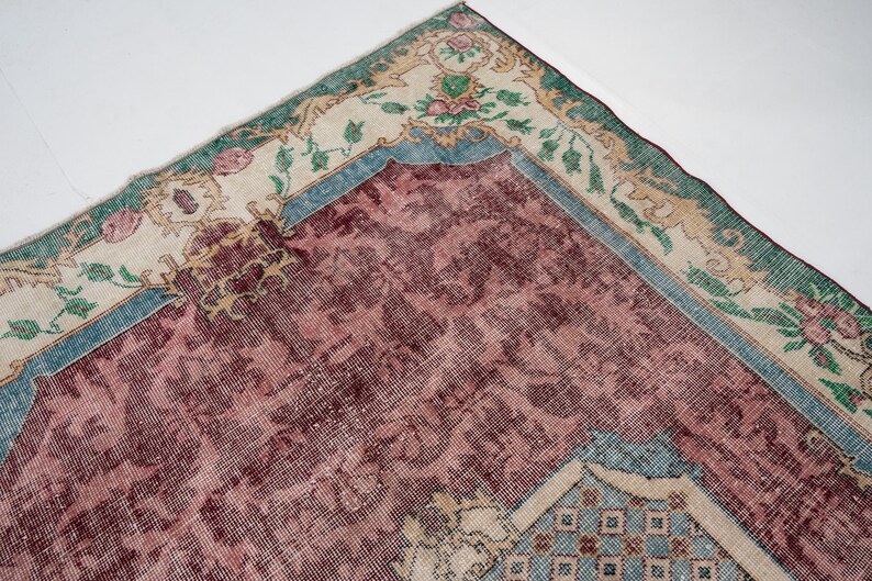 May include: An antique rug with a distressed, vintage appearance. The rug features a central field of deep burgundy, framed by a border of cream, teal, and green. Floral and geometric patterns adorn the rug's surface, creating a classic, timeless aesthetic.