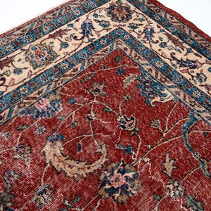 May include: Close-up of a section of a vintage rug with a detailed floral pattern. The rug features a deep red central field with intricate designs in blue, beige, and pink. The border has a contrasting beige and blue pattern.