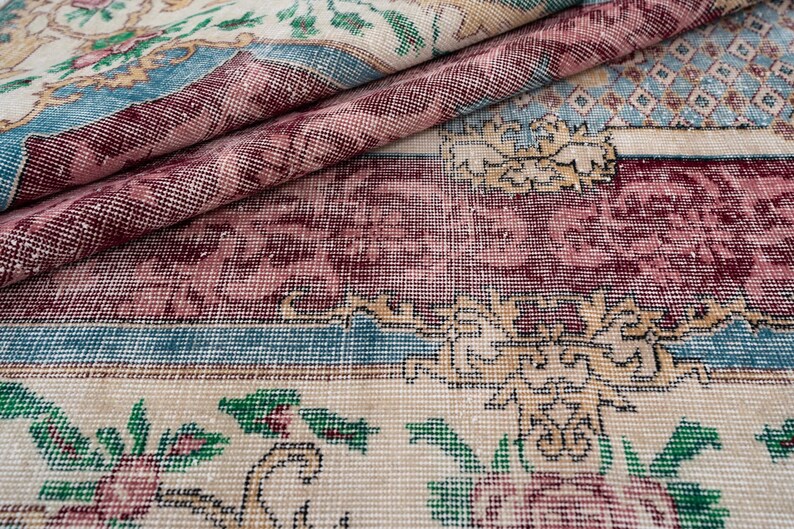 May include: Close-up of a vintage rug with a distressed appearance. The rug features a complex floral pattern in shades of burgundy, teal, cream, and green. The design includes ornate borders and a faded, textured look, suggesting age and use.
