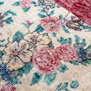 Pode incluir: Close-up de um tapete vintage com um padr&atilde;o floral. O tapete apresenta um fundo creme com flores grandes e detalhadas em tons de rosa, roxo e azul, acentuadas por folhas verdes. Uma se&ccedil;&atilde;o bord&ocirc; &eacute; vis&iacute;vel.