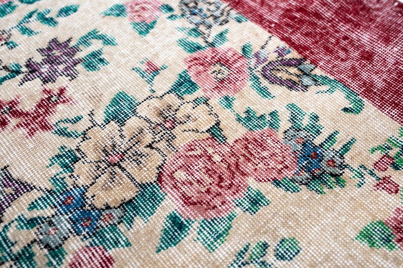 Pode incluir: Close-up de um tapete vintage com um padr&atilde;o floral. O tapete apresenta um fundo creme com flores grandes e detalhadas em tons de rosa, roxo e azul, acentuadas por folhas verdes. Uma se&ccedil;&atilde;o bord&ocirc; &eacute; vis&iacute;vel.