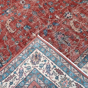 May include: A detailed shot of a red and beige area rug with an intricate floral pattern. The rug features a central field of red with floral motifs in blue, beige, and green. The border is a contrasting beige with a teal and red geometric design.