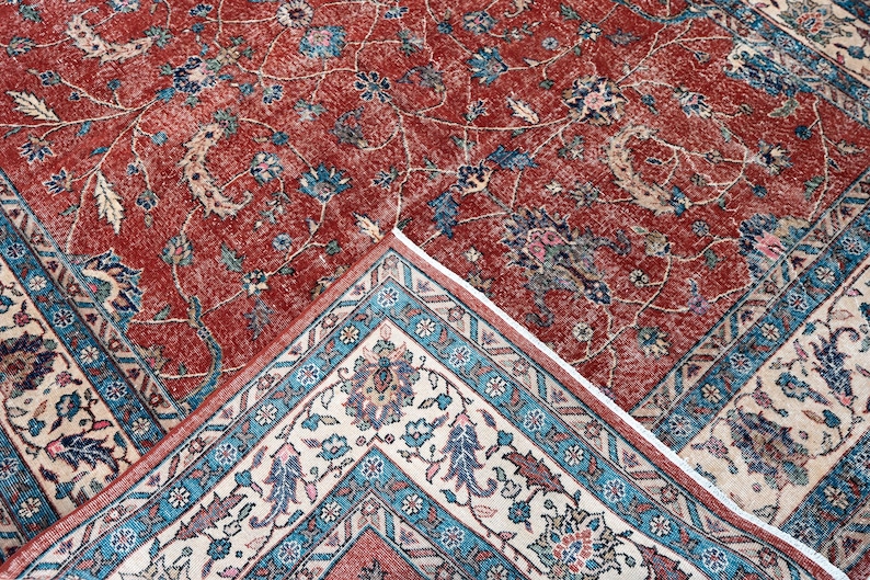 May include: A detailed shot of a red and beige area rug with an intricate floral pattern. The rug features a central field of red with floral motifs in blue, beige, and green. The border is a contrasting beige with a teal and red geometric design.