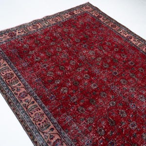 K&ouml;nnte beinhalten: Ein rechteckiger, verwaschen roter Teppich mit floralem Muster. Der Teppich hat ein verblasstes Aussehen, mit einer dunkleren roten Mitte und einem helleren roten Rand. Der Rand weist ein Blumendesign in Rosa-, Gr&uuml;n- und Graut&ouml;nen auf.