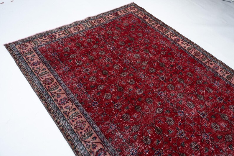 K&ouml;nnte beinhalten: Ein rechteckiger, verwaschen roter Teppich mit floralem Muster. Der Teppich hat ein verblasstes Aussehen, mit einer dunkleren roten Mitte und einem helleren roten Rand. Der Rand weist ein Blumendesign in Rosa-, Gr&uuml;n- und Graut&ouml;nen auf.