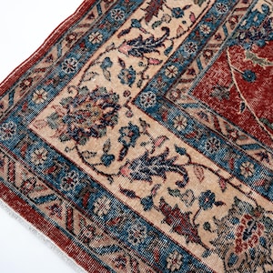 May include: Close-up of a section of a vintage rug with a detailed floral pattern. The rug features a red field with a cream-colored border and blue accents. The design includes stylized flowers and leaves.