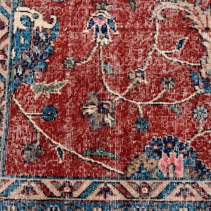 May include: Close-up of a section of a vintage rug with a distressed, faded appearance. The rug features a red central field with floral and vine patterns in blue, green, and beige. A blue border with geometric designs frames the central field.