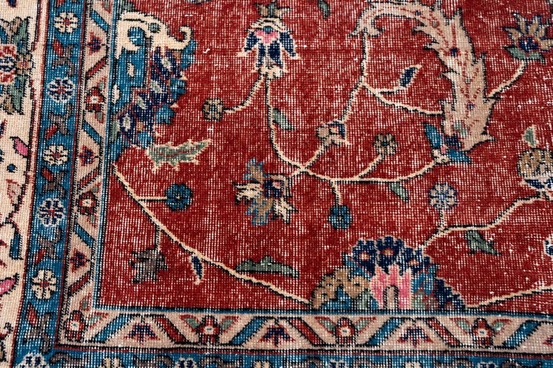 May include: Close-up of a section of a vintage rug with a distressed, faded appearance. The rug features a red central field with floral and vine patterns in blue, green, and beige. A blue border with geometric designs frames the central field.