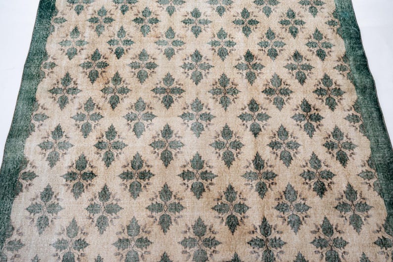 May include: A rectangular area rug featuring a repeating diamond-shaped floral pattern in dark green against a light beige background. The rug is framed by a solid dark green border that runs along the edges, creating a distinct contrast.