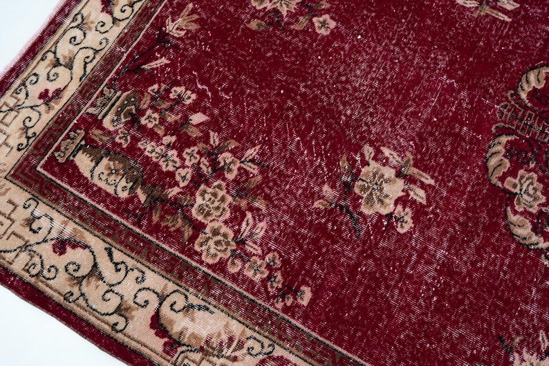 May include: Close-up of a section of a vintage red rug with floral patterns. The rug features a deep red field with faded areas and beige borders with intricate designs. Beige and brown floral motifs are scattered across the rug.