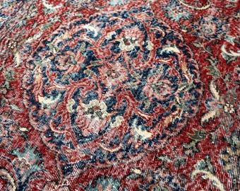 May include: Close-up of a distressed, vintage rug with a central medallion design. The rug features a rich color palette of red, navy blue, and cream, with intricate floral and arabesque patterns. The rug's texture is visible, suggesting a woven construction.