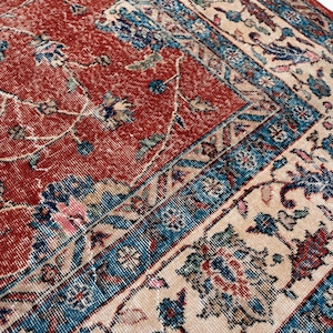 May include: Close-up of a vintage rug with a distressed appearance. The rug features a red central field with floral patterns and a cream border with blue and red accents. The design is intricate and detailed.