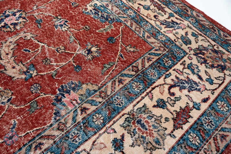 May include: Close-up of a vintage rug with a distressed appearance. The rug features a red central field with floral patterns and a cream border with blue and red accents. The design is intricate and detailed.