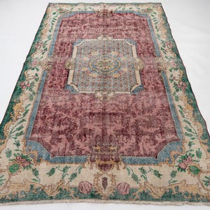 May include: A rectangular, vintage-style rug with a central maroon field and a decorative border. The border features floral and geometric patterns in shades of green, blue, and beige. The rug's design includes a central medallion and corner accents.
