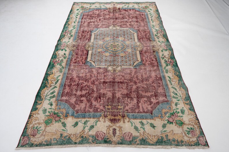May include: A rectangular, vintage-style rug with a central maroon field and a decorative border. The border features floral and geometric patterns in shades of green, blue, and beige. The rug's design includes a central medallion and corner accents.