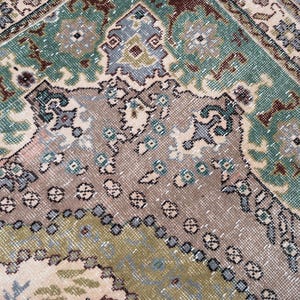 May include: A woven rug featuring a complex geometric and floral pattern in shades of sage green, muted brown, beige, and soft blue. The design includes intricate borders and central motifs with a textured, knotted surface and visible fiber details.