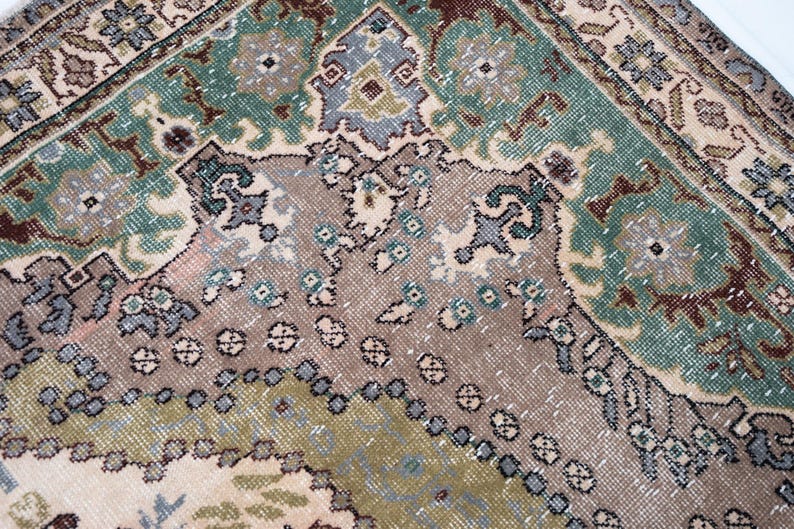 May include: A woven rug featuring a complex geometric and floral pattern in shades of sage green, muted brown, beige, and soft blue. The design includes intricate borders and central motifs with a textured, knotted surface and visible fiber details.