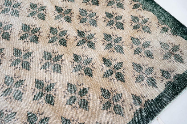 May include: A rectangular area rug featuring a repeating pattern of dark green, four-leaf floral motifs set against a light beige background. The rug has a wide, solid dark green border along the edge, showcasing a woven texture throughout the surface.