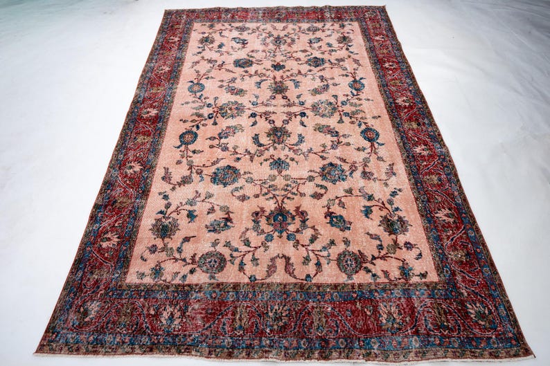 Pu&ograve; includere: Un tappeto rettangolare in stile vintage, con un campo centrale color pesca ornato da motivi floreali blu e turchese. Il bordo &egrave; rosso intenso con disegni intricati. Il tappeto, probabilmente in lana, &egrave; adatto per un soggiorno o una camera da letto.