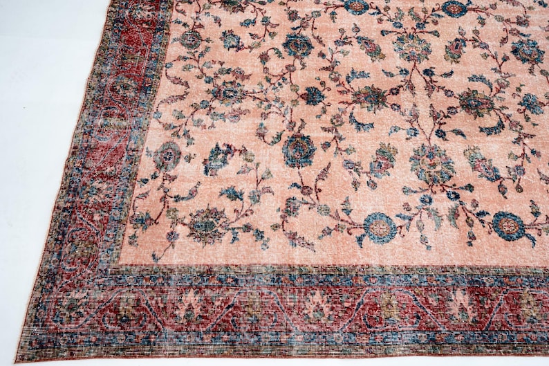 Pu&ograve; includere: Un grande tappeto rettangolare con un'estetica vintage sbiadita. Il tappeto presenta un campo centrale color pesca con un motivo floreale nei toni del blu, verde e marrone. Un bordo dettagliato in rosso e blu incornicia il design, aggiungendo un tocco classico.