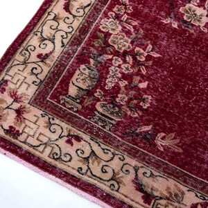 May include: A section of a vintage rug with a deep red background and a beige border. The border features floral and geometric patterns in beige, brown, and red. The rug's design includes floral motifs and vase illustrations.