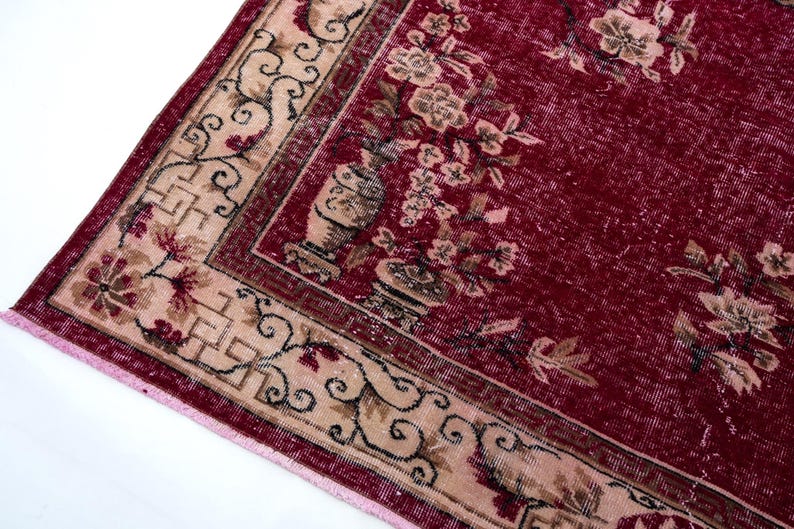 May include: A section of a vintage rug with a deep red background and a beige border. The border features floral and geometric patterns in beige, brown, and red. The rug's design includes floral motifs and vase illustrations.
