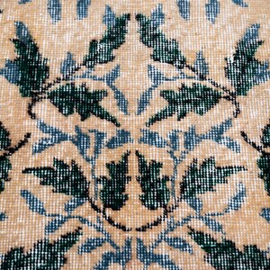 May include: Close-up of a beige rug with a repeating floral pattern in dark green and light blue. The rug has a textured appearance, with the design elements appearing slightly faded.