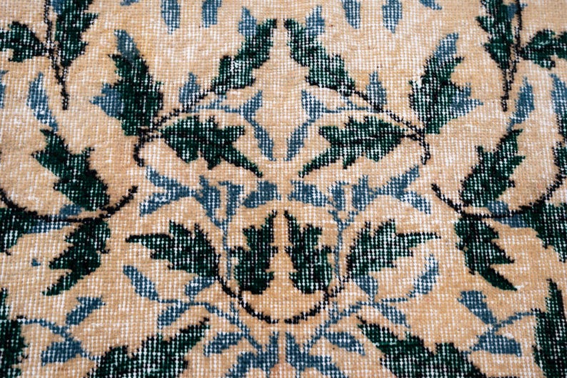 May include: Close-up of a beige rug with a repeating floral pattern in dark green and light blue. The rug has a textured appearance, with the design elements appearing slightly faded.