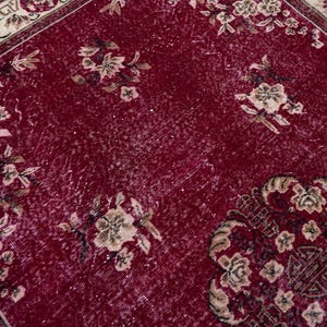 May include: A close-up of a red rug with floral patterns in beige and brown. The rug has a distressed look, with faded areas and a detailed border. The design features multiple floral arrangements.