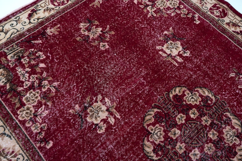 May include: A close-up of a red rug with floral patterns in beige and brown. The rug has a distressed look, with faded areas and a detailed border. The design features multiple floral arrangements.