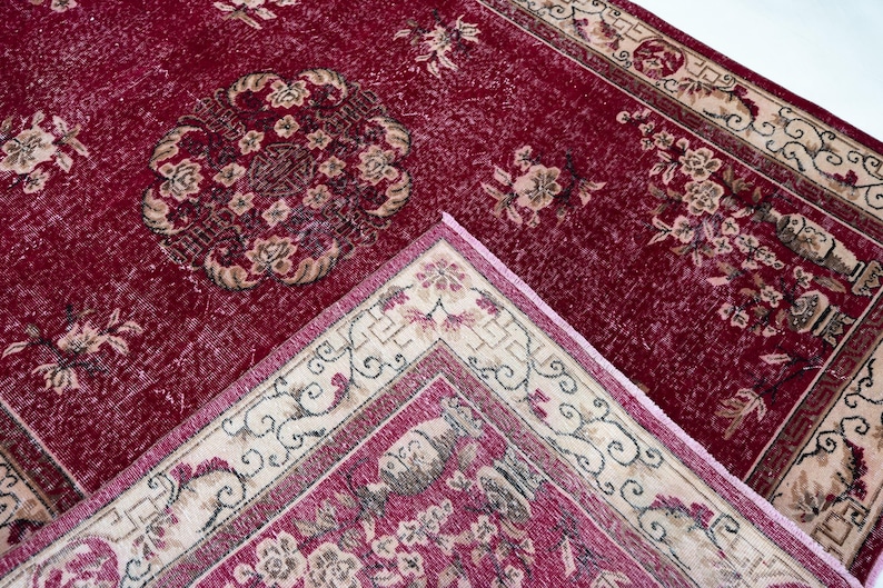 May include: A close-up of a red vintage rug with floral patterns. The rug has a central medallion design and a cream-colored border with floral and scroll motifs. The rug is made of a soft, textured material.