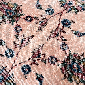Pu&ograve; includere: Primo piano di un tappeto vintage con motivo floreale. Il tappeto presenta uno sfondo color pesca con un aspetto invecchiato. Il design floreale include accenti verde acqua, blu navy e bordeaux, creando un'estetica classica e invecchiata.