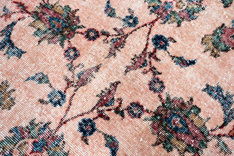 Pu&ograve; includere: Primo piano di un tappeto vintage con motivo floreale. Il tappeto presenta uno sfondo color pesca con un aspetto invecchiato. Il design floreale include accenti verde acqua, blu navy e bordeaux, creando un'estetica classica e invecchiata.