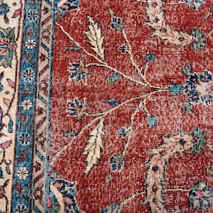 May include: Close-up of a section of a vintage rug with a distressed, faded appearance. The rug features a red background with floral patterns in blue, cream, and green. The border has a cream background with blue and floral accents.