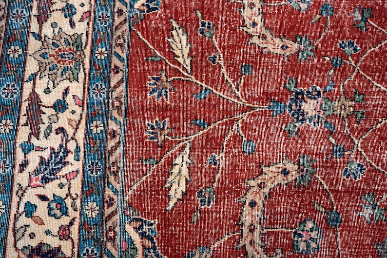 May include: Close-up of a section of a vintage rug with a distressed, faded appearance. The rug features a red background with floral patterns in blue, cream, and green. The border has a cream background with blue and floral accents.