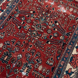 May include: A rectangular area rug featuring a deep red field with intricate floral and geometric patterns in shades of blue, cream, and black. The border displays a repeating floral motif on a dark blue background with cream accents.