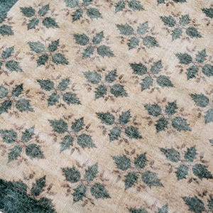 May include: A rectangular area rug featuring a repeating pattern of dark teal four-petaled floral motifs set against a light beige background. The rug includes a solid dark teal border along the edge, showcasing a woven texture throughout.
