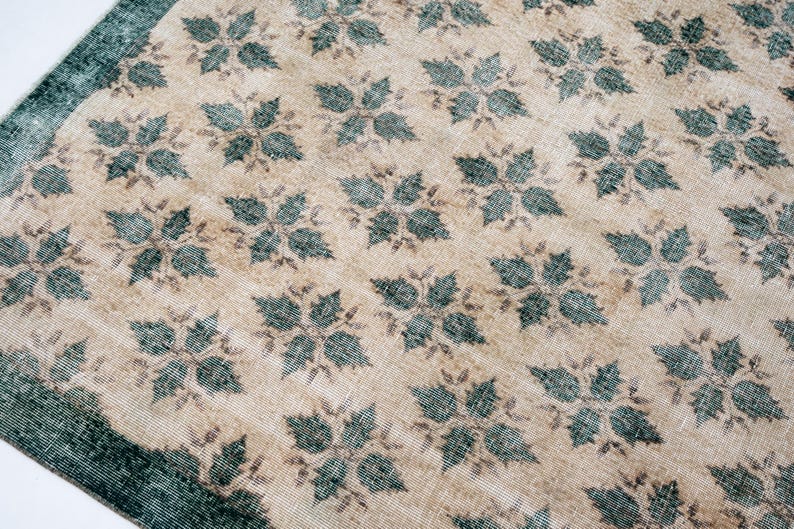 May include: A rectangular area rug featuring a repeating pattern of dark teal four-petaled floral motifs set against a light beige background. The rug includes a solid dark teal border along the edge, showcasing a woven texture throughout.