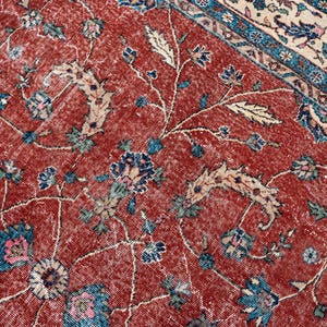 May include: Close-up of a distressed, vintage rug with a red background and floral patterns. The rug features blue, pink, and cream-colored floral and vine designs. The border has a blue and cream pattern.