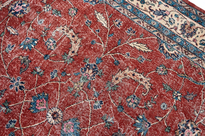 May include: Close-up of a distressed, vintage rug with a red background and floral patterns. The rug features blue, pink, and cream-colored floral and vine designs. The border has a blue and cream pattern.