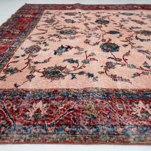 Pu&ograve; includere: Un grande tappeto rettangolare dall'aspetto sbiadito e vintage. Il tappeto presenta un campo centrale color pesca chiaro con motivi floreali blu e verdi. Un ampio bordo rosso e blu incornicia il disegno.