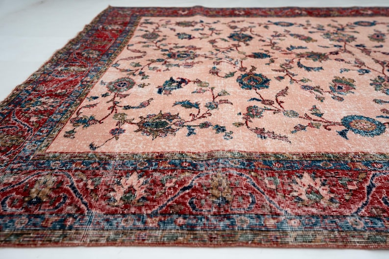 Pu&ograve; includere: Un grande tappeto rettangolare dall'aspetto sbiadito e vintage. Il tappeto presenta un campo centrale color pesca chiaro con motivi floreali blu e verdi. Un ampio bordo rosso e blu incornicia il disegno.