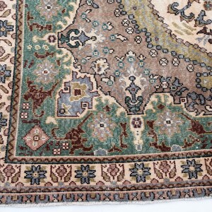 May include: A woven area rug featuring a complex pattern with a central field in shades of brown, green, and cream. The border displays repeating floral motifs in blue, pink, and brown, with intricate geometric shapes throughout the design.