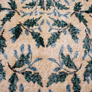 May include: Close-up of a beige rug with a repeating pattern of green and blue leaves. The leaves are arranged in a symmetrical design across the rug's surface, creating a textured appearance.
