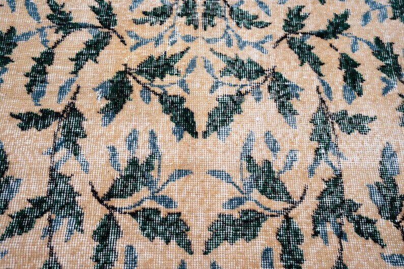 May include: Close-up of a beige rug with a repeating pattern of green and blue leaves. The leaves are arranged in a symmetrical design across the rug's surface, creating a textured appearance.